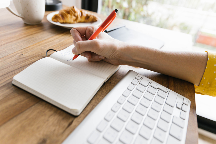 Unrecognizable crop female entrepreneur writing in notebook with pen while sitting at wooden desktop with keyboard cup of coffee and croissant on desk on sunny day